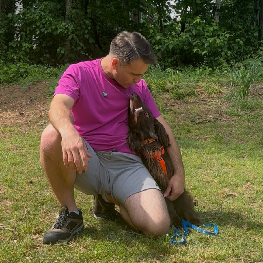 Joel Harrison kneeling outdoors in the grass with a brown dog leaning into him.